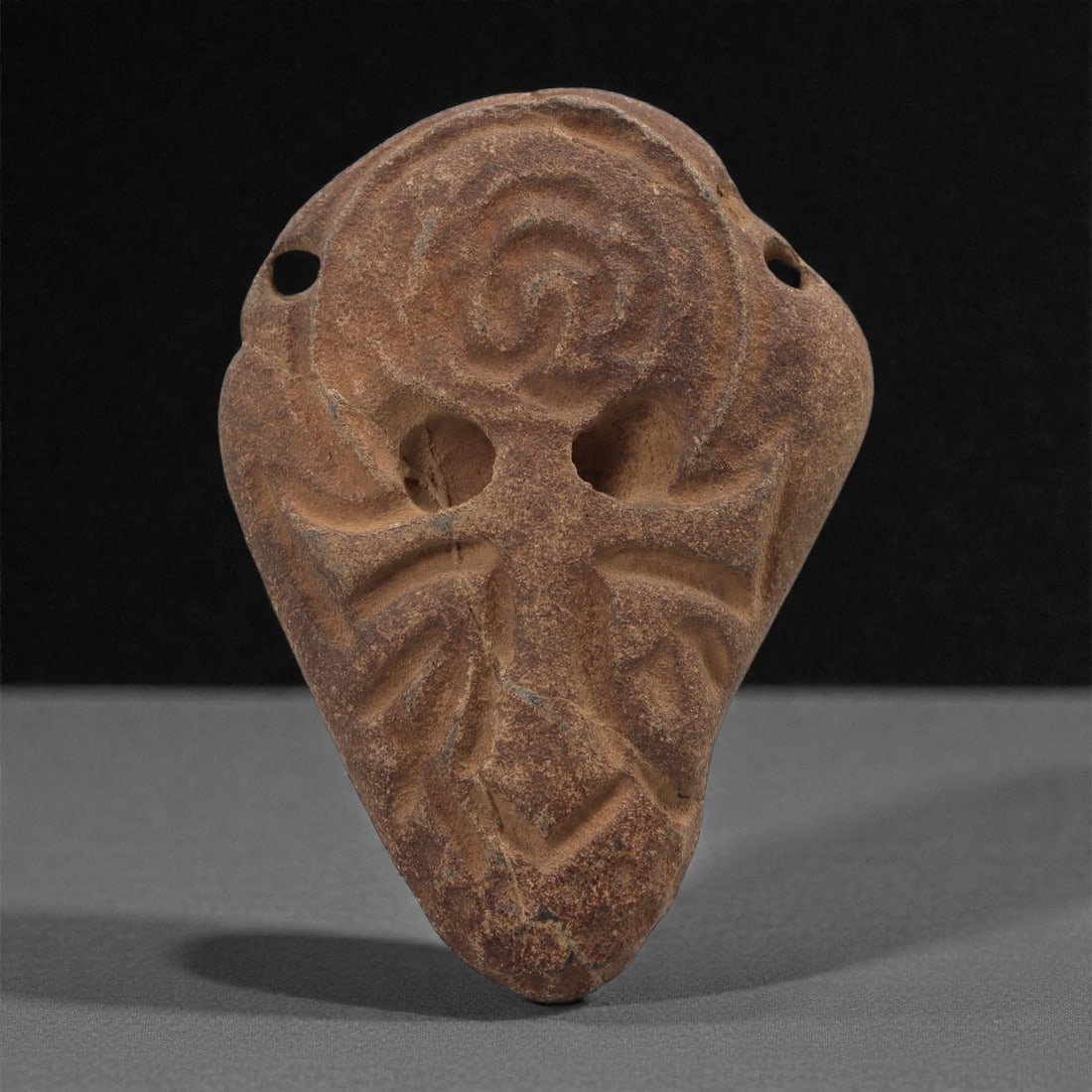 A Byzantine Stone Amulet with Cross and Spiral Motif, 10th-12th Century, 80B: Carved from a reddish-brown stone, the amulet is shaped as a tapering triangular plaque with rounded shoulders. The upper field contains a recessed spiral enclosed within a circular border, while two