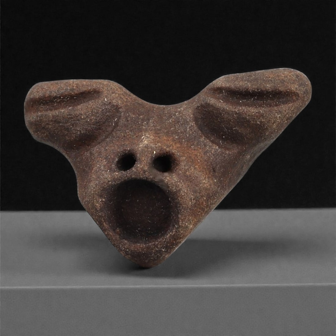 A Mediterranean Stone Maskette, 12th-17th Century, 25AD: Carved from a dark brown stone with a fine granular surface, the maskette is shaped in an unusual triangular configuration with two broad projecting upper extensions. The eyes are rendered as recessed