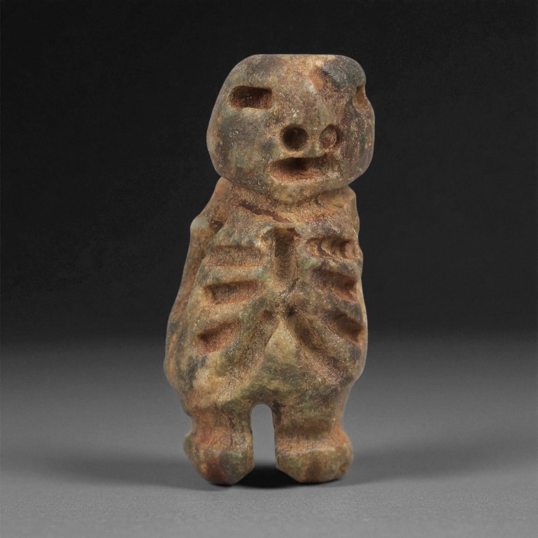 A Taino Carved Stone Idol, Late Ceramic Age, circa 1200-1500 CE, 89LD: Carved from mottled brown-green stone, the figure is rendered in a standing posture with short legs and a rounded head. The facial features consist of drilled circular eyes and mouth, with recessed ar