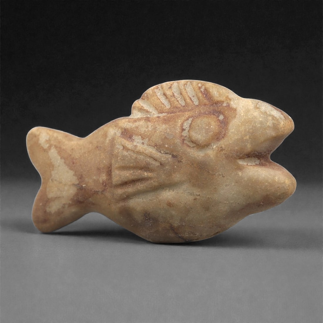 An Egyptian Stone Fish Figurine, New Kingdom, Circa 1550-1070 BCE, 80LF: Carved from a light brown stone with surface accretions, the object is shaped as a stylized fish with a broad body, a forked tail, and an open mouth. Incised lines define the gill, fins, and dorsal el