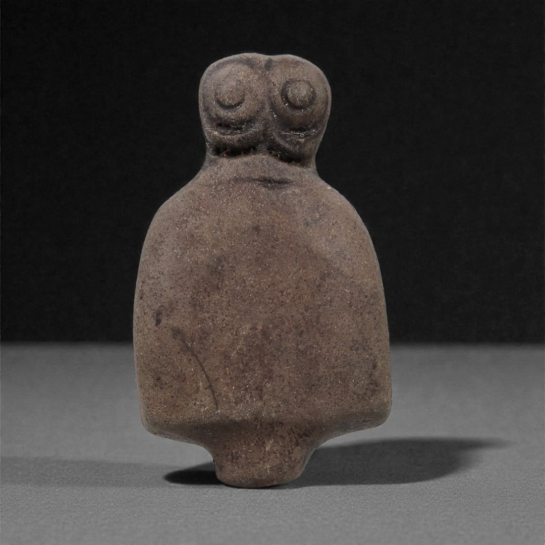 A Near Eastern Stone Eye Idol, 4th-3rd Millennium BCE, 48LH: Carved from a dark brown stone with a finely granular surface, the figure is shaped with a broad, rounded lower body tapering to a short projection at the base. The upper section forms a narrow neck s