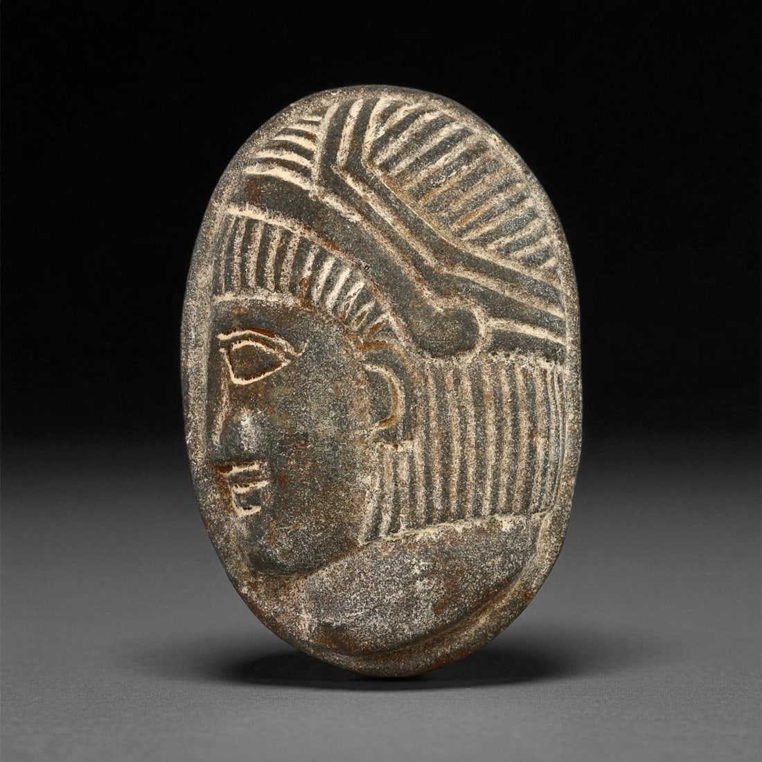 A Sasanian Stone Intaglio Plaque, 3rd-7th Century CE, 5.6cm, 67ES: The stone plaque is shaped with a convex surface bearing an incised profile head. The facial features are rendered with linear grooves defining the eye, brow, nose, and mouth, while additional incisio