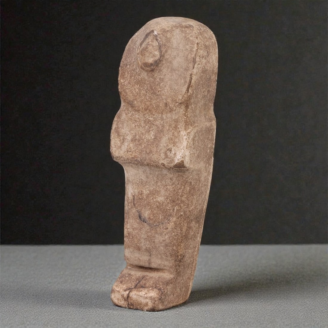 An Egyptian Revival Stone Mummiform Figure, 50R: Carved from light brown to buff stone with visible natural veining, inclusions, and areas of surface abrasion, the object represents a mummiform standing figure rendered in a simplified, frontal manne