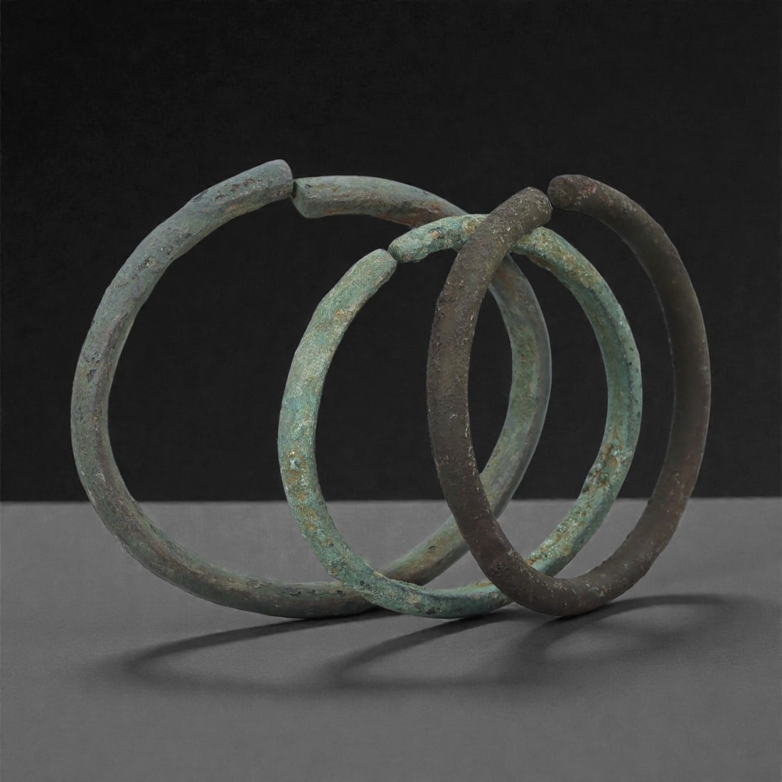 Roman Bronze Bracelets, 1st-3rd Century CE, 4H: Cast in bronze with open-ended circular forms, the group comprises three bracelets of rounded profile showing burial-derived patination ranging from pale green to dark brown. The surfaces display mino