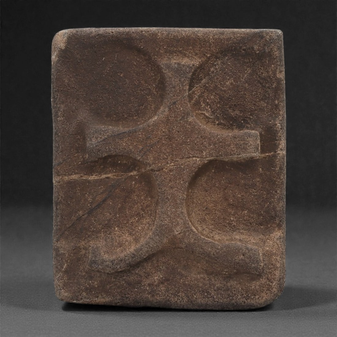 A Byzantine Stone Relief Plaque with Cross Form, 10th-12th Century, 65A: Carved from a dark brown stone with a fine, even surface, the plaque bears a shallow relief of a cross composed of four rounded, concave terminals joined by short, straight arms. The recessed quadrant