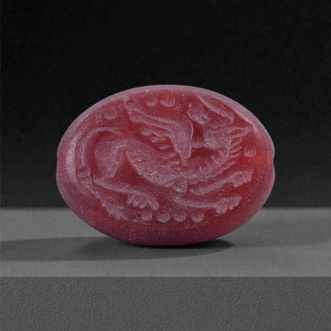A Sasanian Red Stone Intaglio with Winged Hybrid Creature, 3rd-7th Century CE, 98J: Carved from an opaque red stone, the oval intaglio bears an incised depiction of a winged composite creature shown in profile, its body defined by shallow linear cutting. The figure displays a feline-