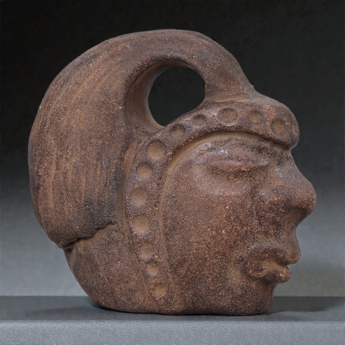 A Mesoamerican Stone Head Pendant, Pre-Columbian Period, 31R: Carved from a single piece of fine-grained brown stone, the object represents a stylized human head shown in profile. The facial features are modeled in shallow relief, with a gently recessed almond-s