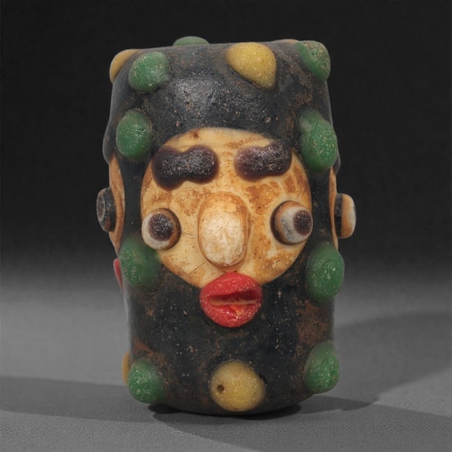 A European Glass Face Bead, 19th-20th Century, 69A: Formed in multicolored glass with an applied mask-like face, the bead is constructed around a cylindrical core with added elements in contrasting hues to create the eyes, brows, nose, and mouth. Addit