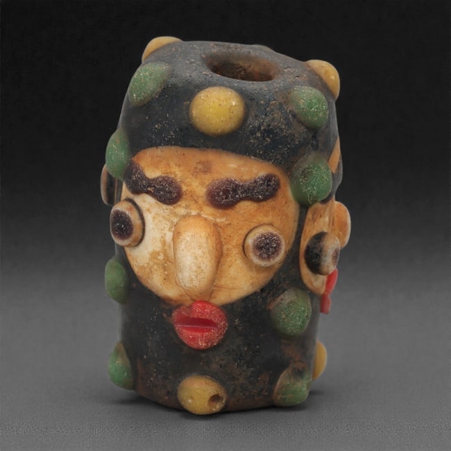 A European Glass Face Bead, 19th-20th Century, 68A: Formed in multicolored glass with an applied mask-like face, the bead is constructed around a cylindrical core with added elements in contrasting hues to create the eyes, brows, nose, and mouth. Addit