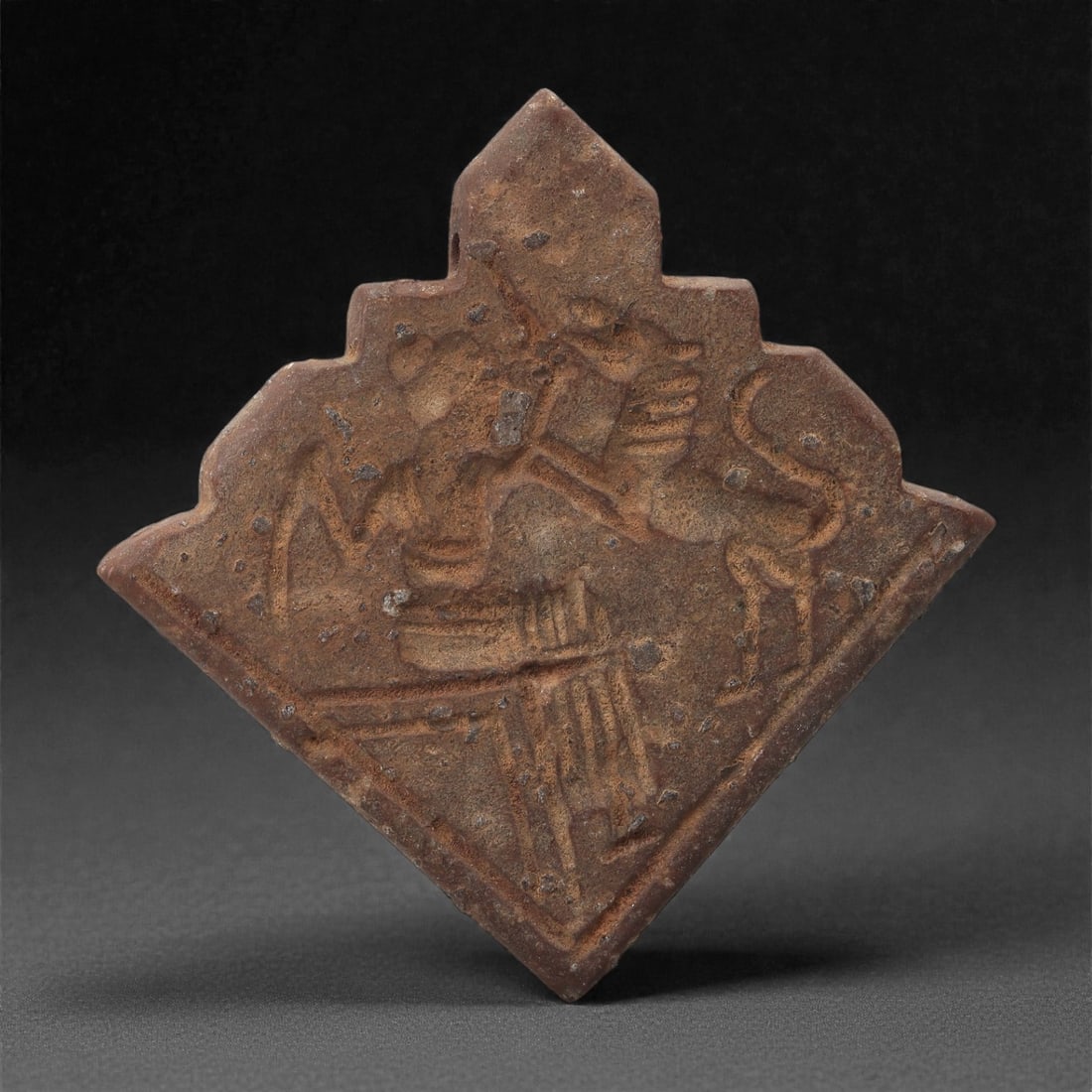 A Sasanian Stone Plaque Amulet, 3rd-7th Century CE, 45LR: Carved from a fine-grained stone with brown archaeological surface accretions, the amulet is configured as a diamond-shaped plaque with stepped upper edges. The face is worked in shallow relief depict