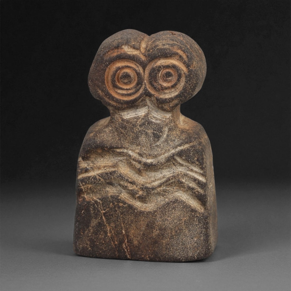 A Near Eastern Stone Eye Idol, 4th-3rd Millennium BCE, 92ZA: Carved from a dark brown stone with lighter veining and surface accretions, the figure is shaped with a rounded head bearing two recessed circular eyes formed by concentric grooves. The shoulders slop