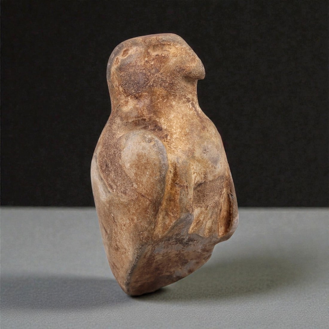 A Near Eastern Stone Bird Amulet, Circa 1st Millennium BCE, 14ZF: Carved from light brown stone with darker mineral inclusions and natural veining, the object represents a stylized bird rendered in the round. The form is compact and vertically oriented, with the hea