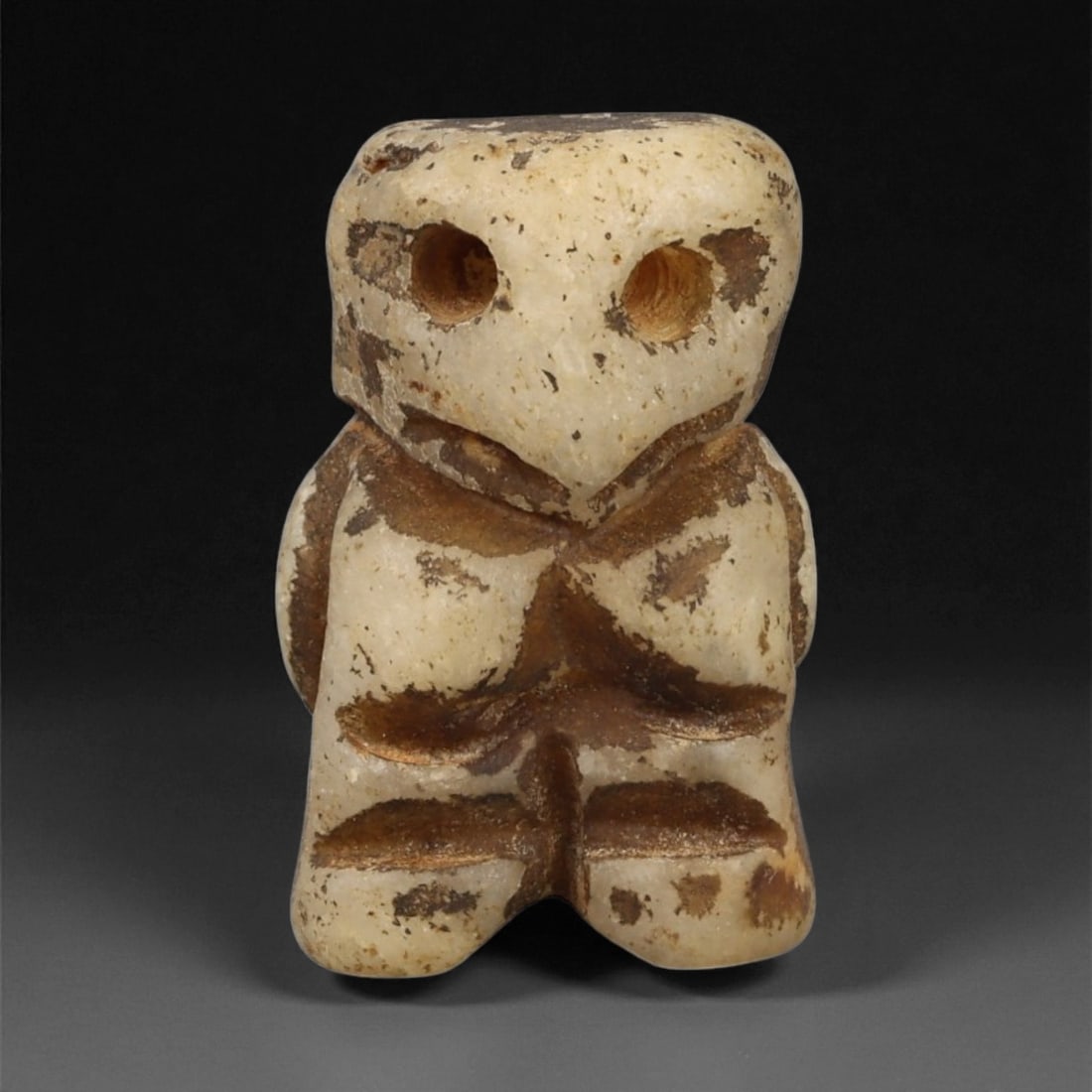 A Taino Carved Stone Idol, Late Pre-Columbian Period, Circa 1000-1500 CE, 69LA: Carved from a pale stone with brown surface deposits, the figure is modeled with a squared head and compact body. The face bears two circular perforations for the eyes, a pronounced central ridge form