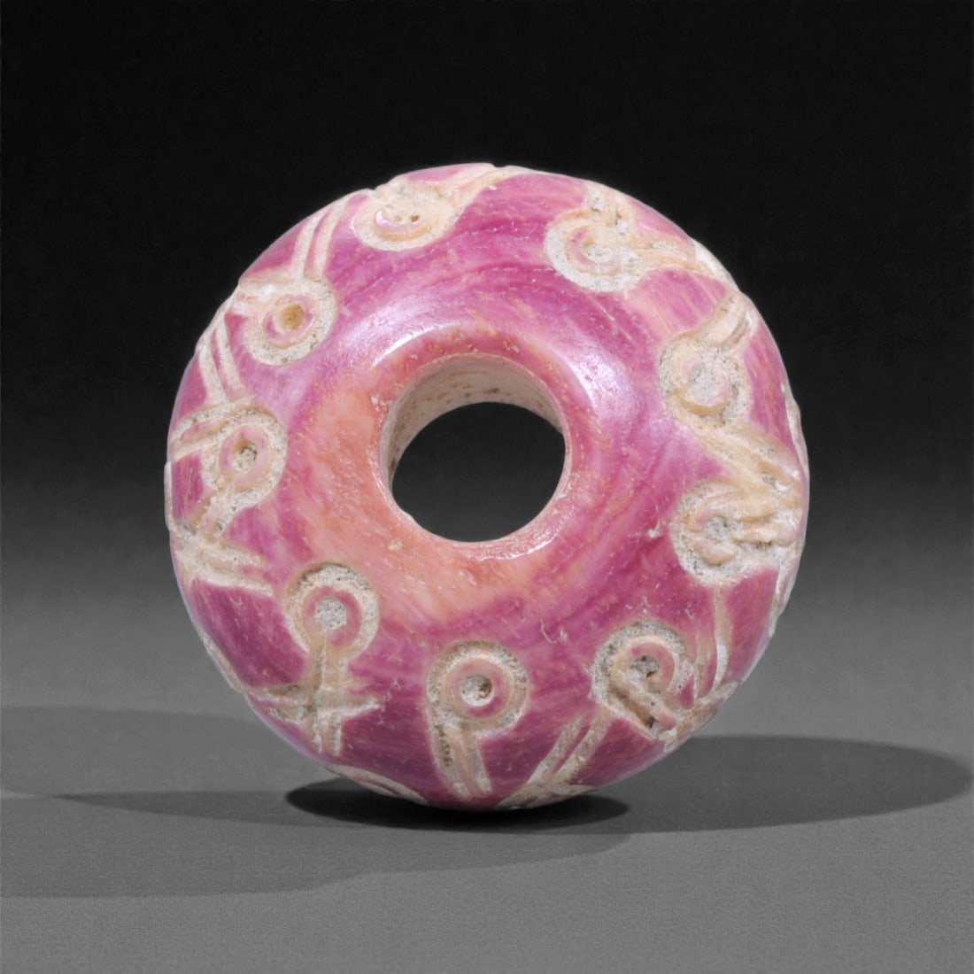 A Near Eastern Bone Spindle Whorl, 1st Millennium BCE, 91D: Carved from a pink-toned bone exhibiting natural striations, the whorl is shaped into a rounded biconvex form with a central perforation. The exterior surface bears a continuous register of incised co