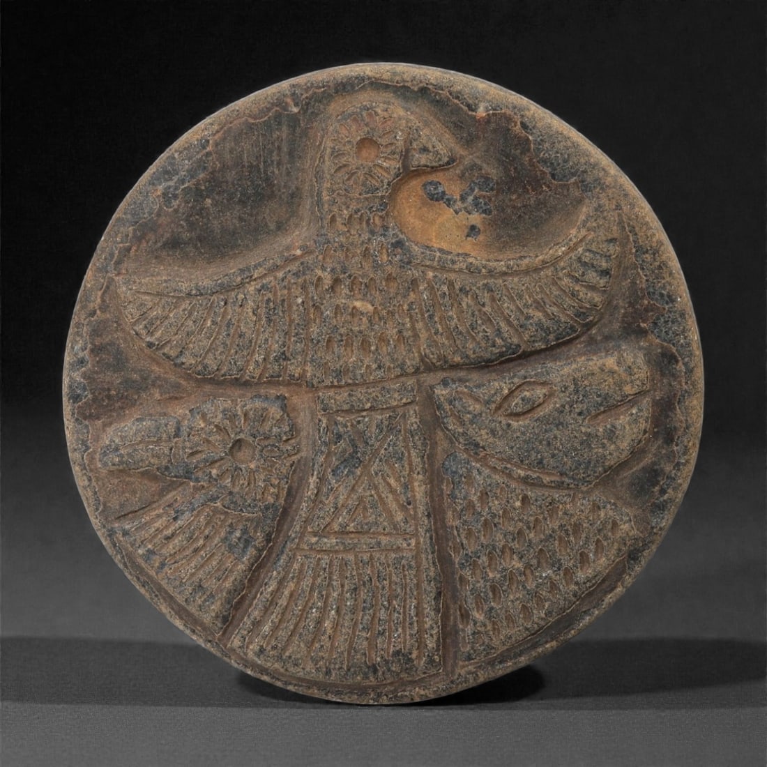 An Indo-Parthian Stone Plaque, 1st-3rd Century CE, 42LE: Carved from a dark brown stone with lighter surface mottling, the circular plaque bears a recessed relief comprising a central standing figure with outstretched, wing-like arms. Flanking the figure ar