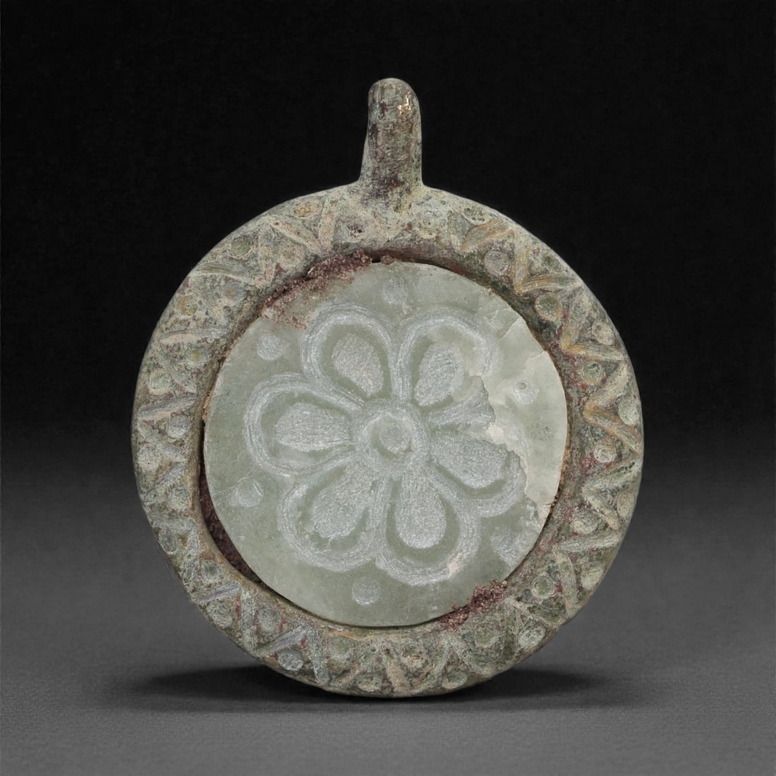 An Islamic Bronze Amulet with Agate Inset, 10th-13th Century CE, 12LP: Cast in bronze with a circular form and an integral suspension loop, the amulet contains a fitted disc of pale agate carved in low relief with a six-petaled rosette centered on a small boss. The bronz