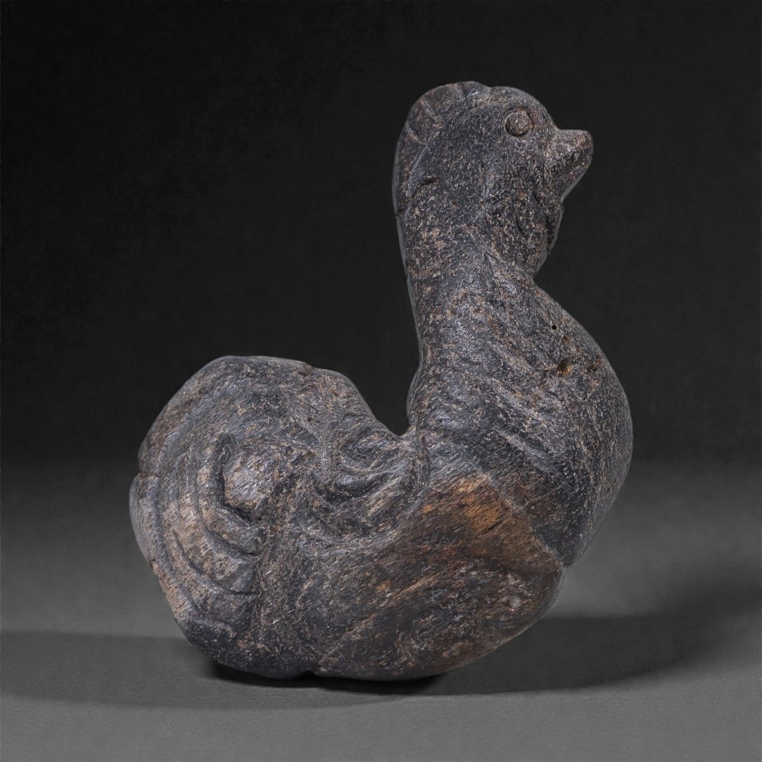 A Near Eastern Stone Avian Effigy, 3rd-2nd Millennium BCE, 14E: Carved from a dark grey stone with fine-grained texture and scattered mineral inclusions, the object represents a standing bird rendered in a compact, volumetric form. The body is rounded and upright,