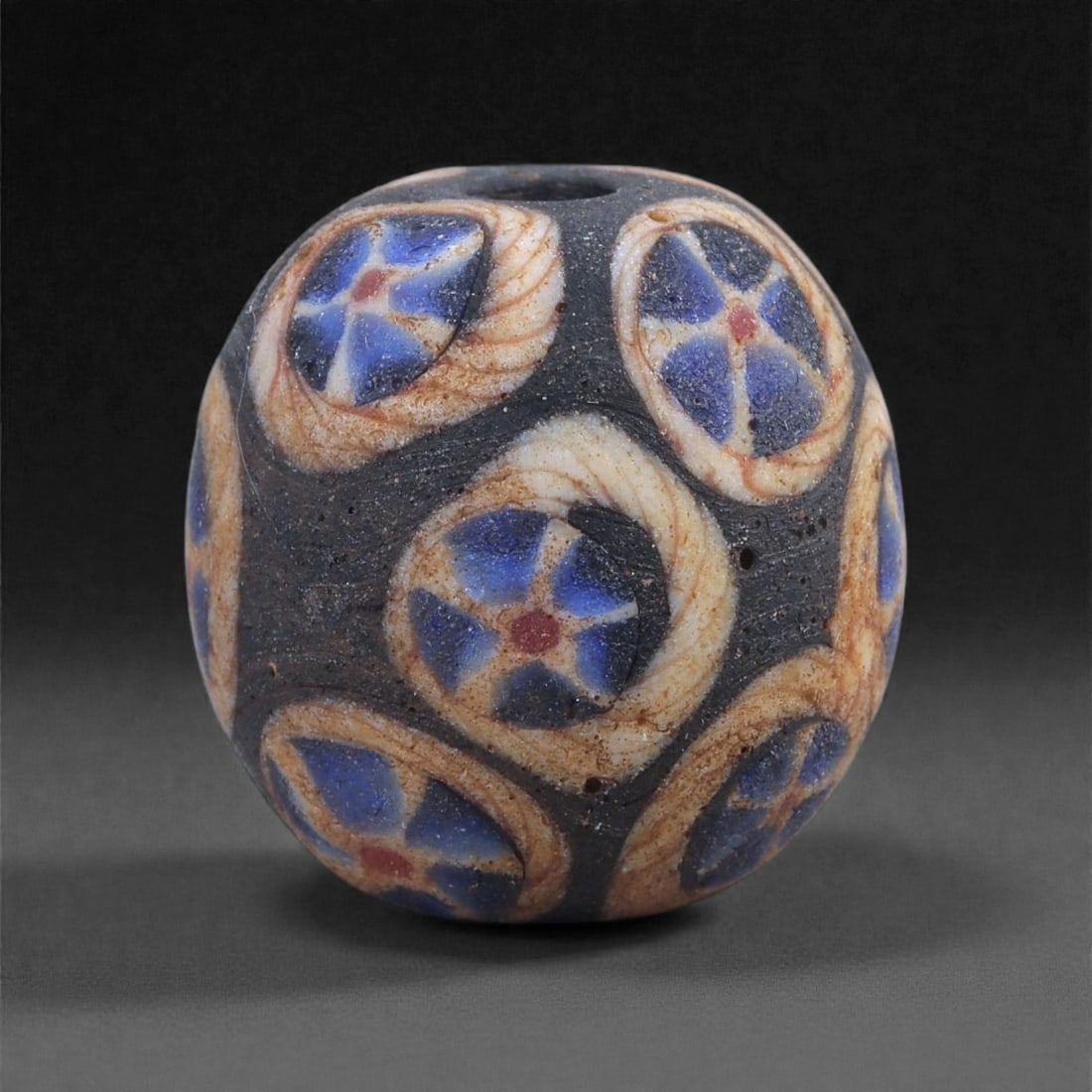 Ancient Egypto-Roman Mosaic Glass Bead - Rosette Cane Sphere, 12J: Formed in dark glass with inlaid mosaic rosette canes, this spherical bead displays a sequence of six-petaled blue rosettes each centered by a red core and encircled by pale spiraling bands. The refin