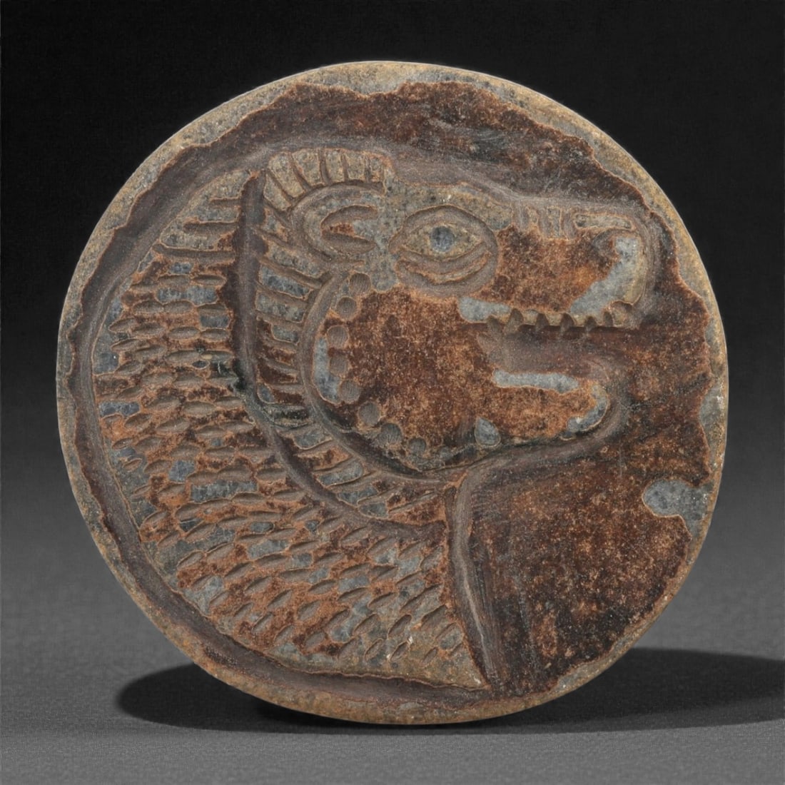 A Near Eastern Stone Plaque with Feline Head, 1st Millennium BCE, 37LE: Carved from a dark brown stone with lighter surface inclusions, the circular plaque bears a recessed relief depicting a feline head in profile. The animal is shown with a rounded eye, open jaw, and a