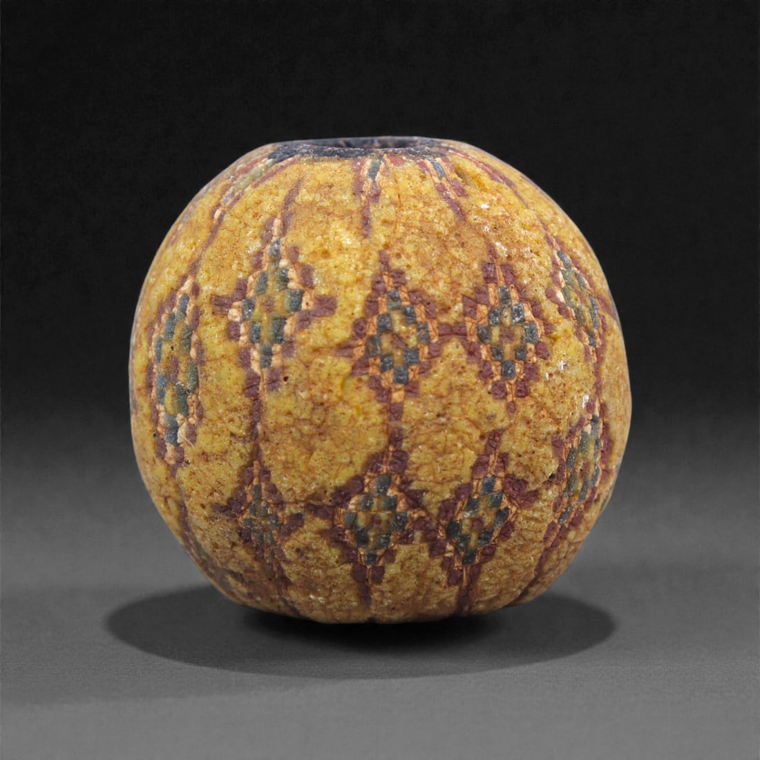 Ancient Egypto-Roman Mosaic Glass Bead - Yellow Ground Millefiori Sphere, 15J: Formed in opaque yellow mosaic glass, this spherical bead incorporates repeating clusters of checkerboard cane slices in green, red, and cream, each arranged within narrow vertical registers that run 