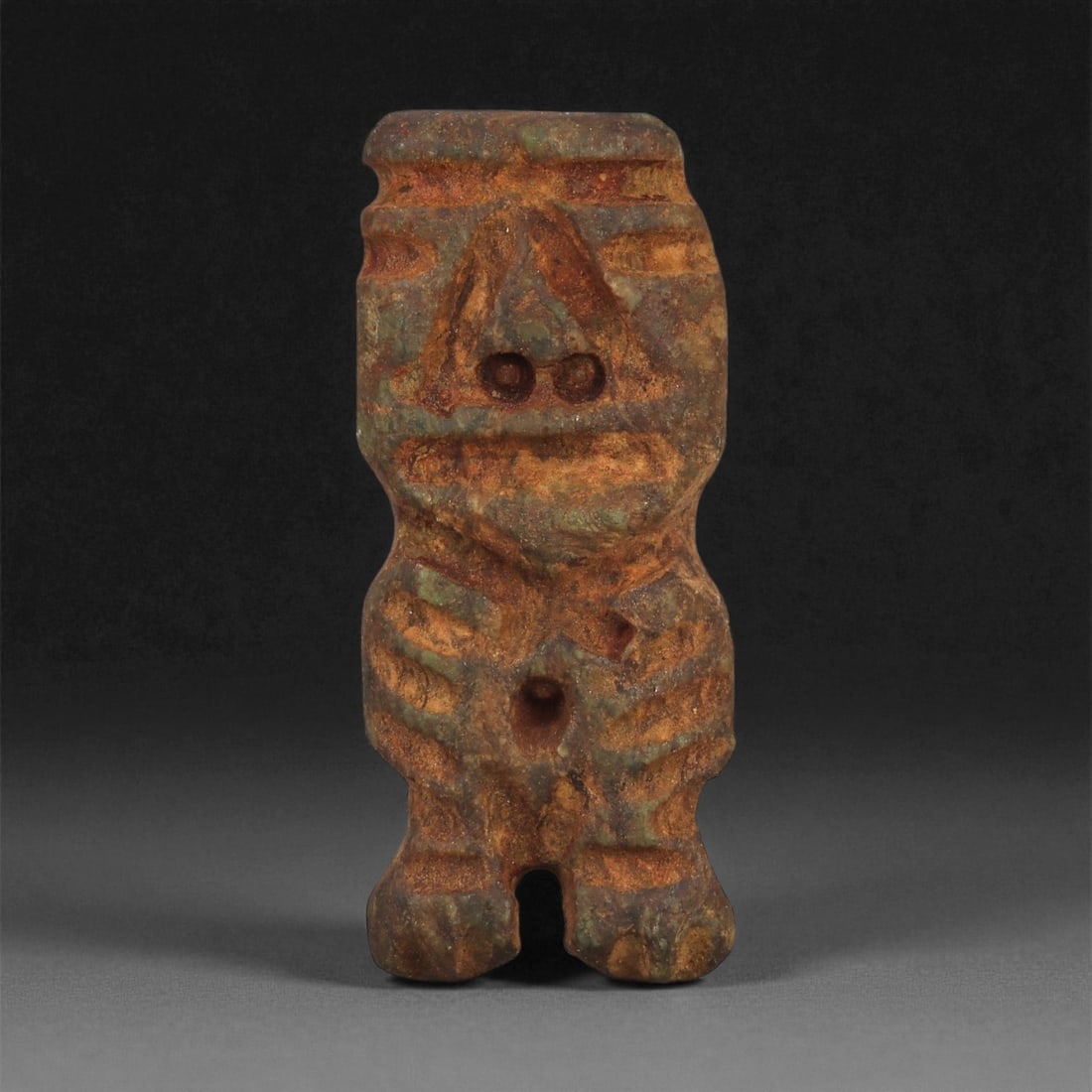 A Taino Carved Stone Idol, Late Pre-Columbian Period, Circa 1000-1500 CE, 97ZA: Carved from mottled brown and green stone, the figure stands upright with short bifurcated legs and a compact torso. The head is defined by recessed circular eyes set beneath a triangular brow, a stra