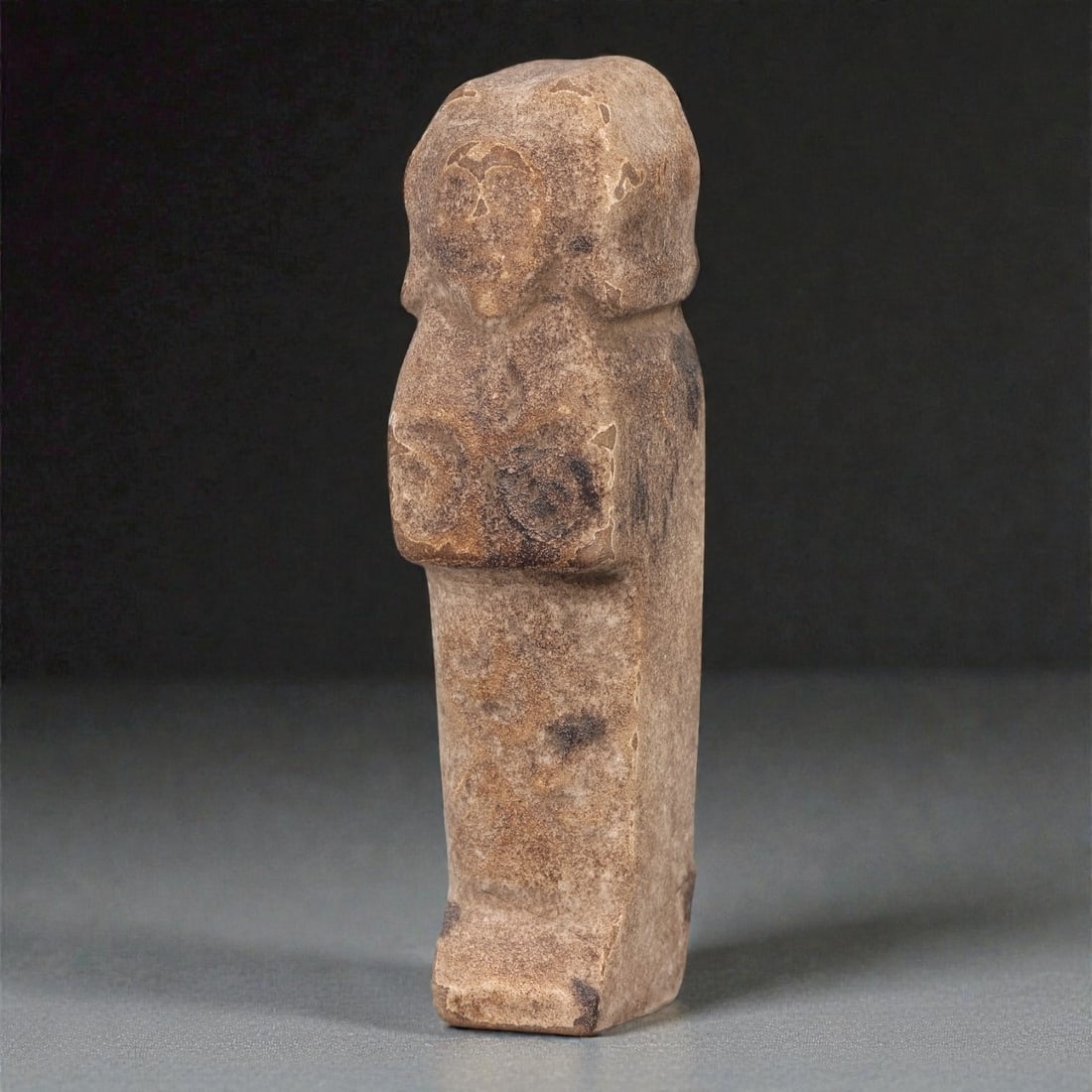 An Egyptian Revival Stone Mummiform Figure, 16R: Carved from light brown to buff stone with scattered darker inclusions and areas of surface abrasion, the object represents a mummiform standing figure rendered in a simplified and frontal manner. The