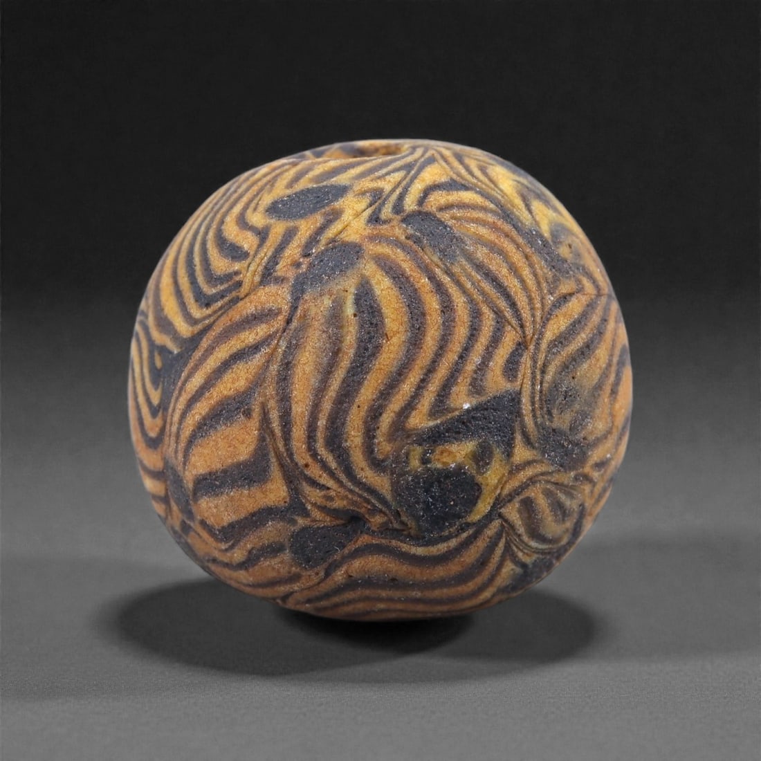 Medieval Islamic Marbled Glass Bead - Banded Swirl Sphere, 9J: Worked in opaque marbled glass, this bead displays an intricate network of undulating dark blue and ochre bands created through the twisting of contrasting colored rods while still molten. The flowing