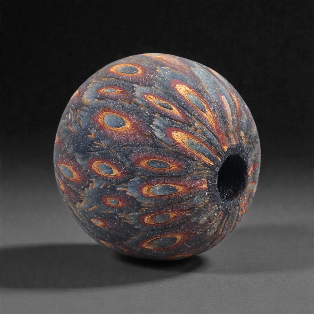 Ancient Egypto-Roman Mosaic Glass Bead - Feathered Eye-Cane Sphere, 20J: Worked in richly colored mosaic glass, this spherical bead displays a continuous surface of elongated eye motifs rendered in layered canes of yellow, red, and blue, each tapering toward the perforatio
