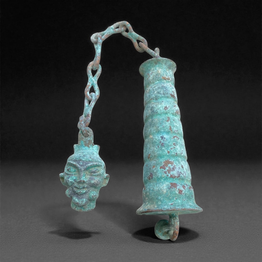 A European Bronze Bell with Mask Terminal, Roman Period, Circa 1st-3rd Century CE, 188g, 11AA: Cast in bronze, the object comprises a small elongated bell suspended from a linked chain terminating in a mask-shaped finial. The bell is cylindrical with a slightly flaring mouth and a looped clappe