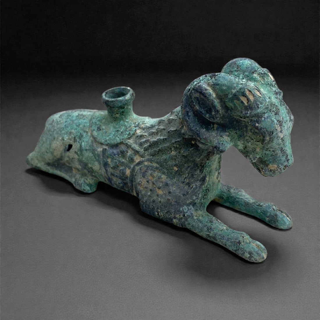 An Achaemenid Bronze Ram-Form Vessel, Circa 5th-4th Century BCE, 210g, 34BB: Cast in bronze in the form of a recumbent ram, the vessel is modeled with the forelegs extended forward and the hindquarters tucked beneath the body. A short cylindrical aperture rises from the upper