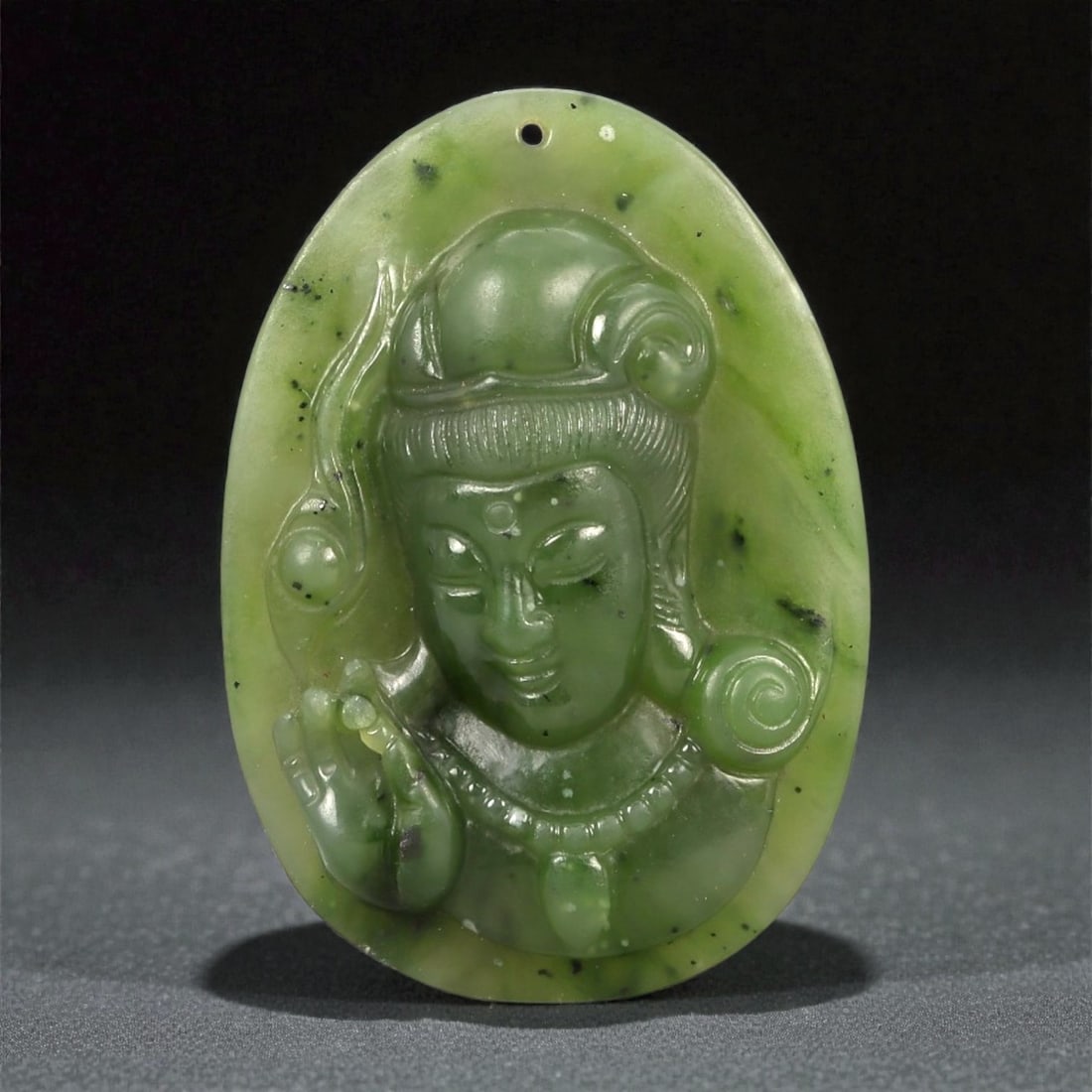A Chinese Nephrite Jade Bodhisattva Pendant, 20th Century, 47FA: Carved from apple-green nephrite jade with areas of translucency and scattered dark mineral inclusions, the pendant is worked in relief to depict the head and upper torso of a bodhisattva, likely Guan