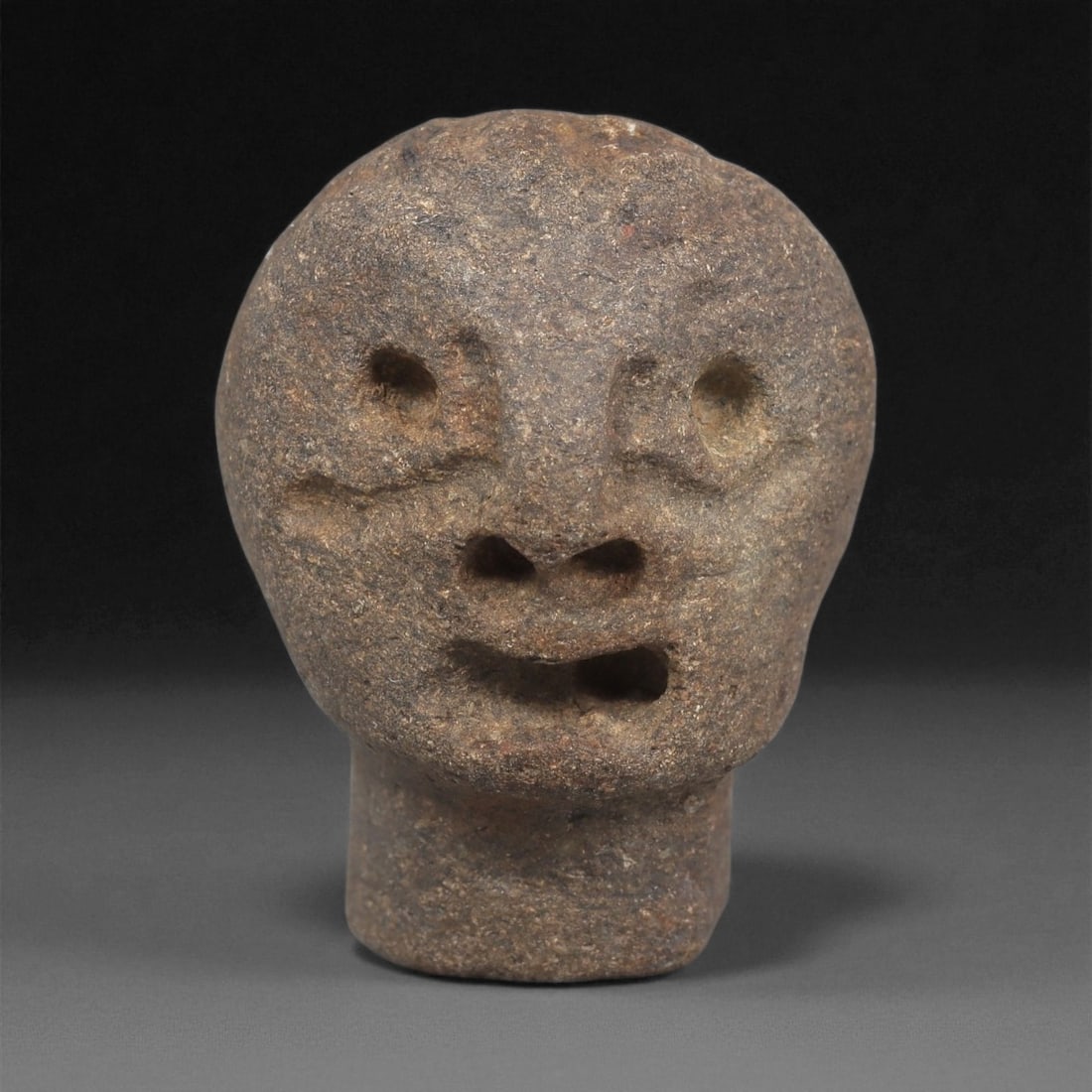 A Mediterranean Stone Maskette, 12th-17th Century, 33ZB: Carved from a coarse-grained brown stone with subtle surface inclusions, the maskette presents a rounded head set atop a short cylindrical neck extension. The facial features are modeled in shallow re