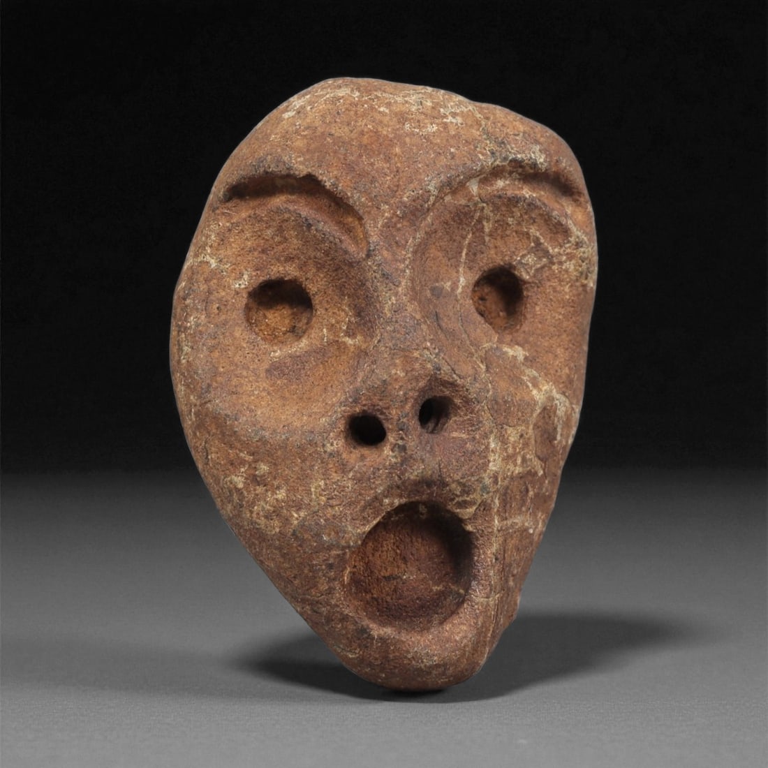 A Mediterranean Stone Maskette, 12th-17th Century, 90ZA: Carved from a reddish-brown stone with lighter surface veining, the maskette presents an elongated oval form tapering toward the chin. The facial features are cut in shallow relief, including large ci