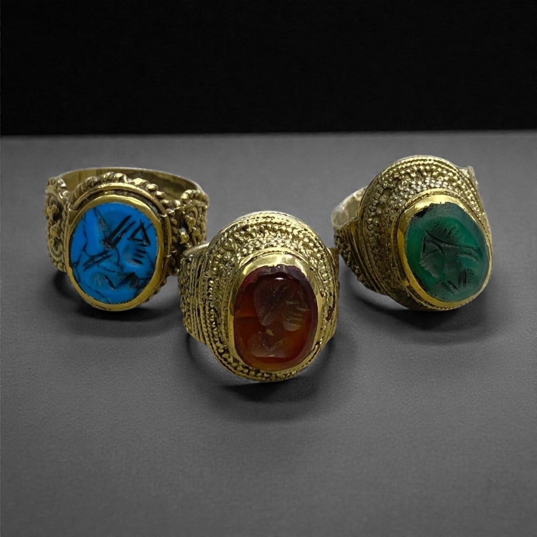 Three Qajar Style Copper Alloy and Glass Rings, 20th Century, US 8-9, 34g, 47AA: Comprising three rings formed from copper alloy, each mounted with an oval glass intaglio carved with figural imagery derived from Qajar-period visual conventions. The bezels are framed by beaded and