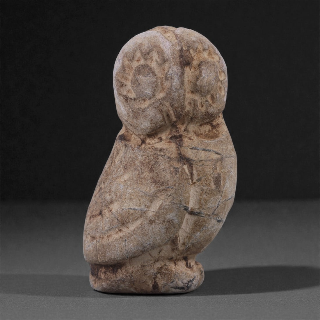 A Near Eastern Stone Zoomorphic Owl Figurine, 3rd-2nd Millennium BCE, 26ZE: Carved from a mottled brown and buff stone with visible natural veining, mineral inclusions, and surface irregularities, this small zoomorphic figurine represents a stylized owl rendered in a compact,