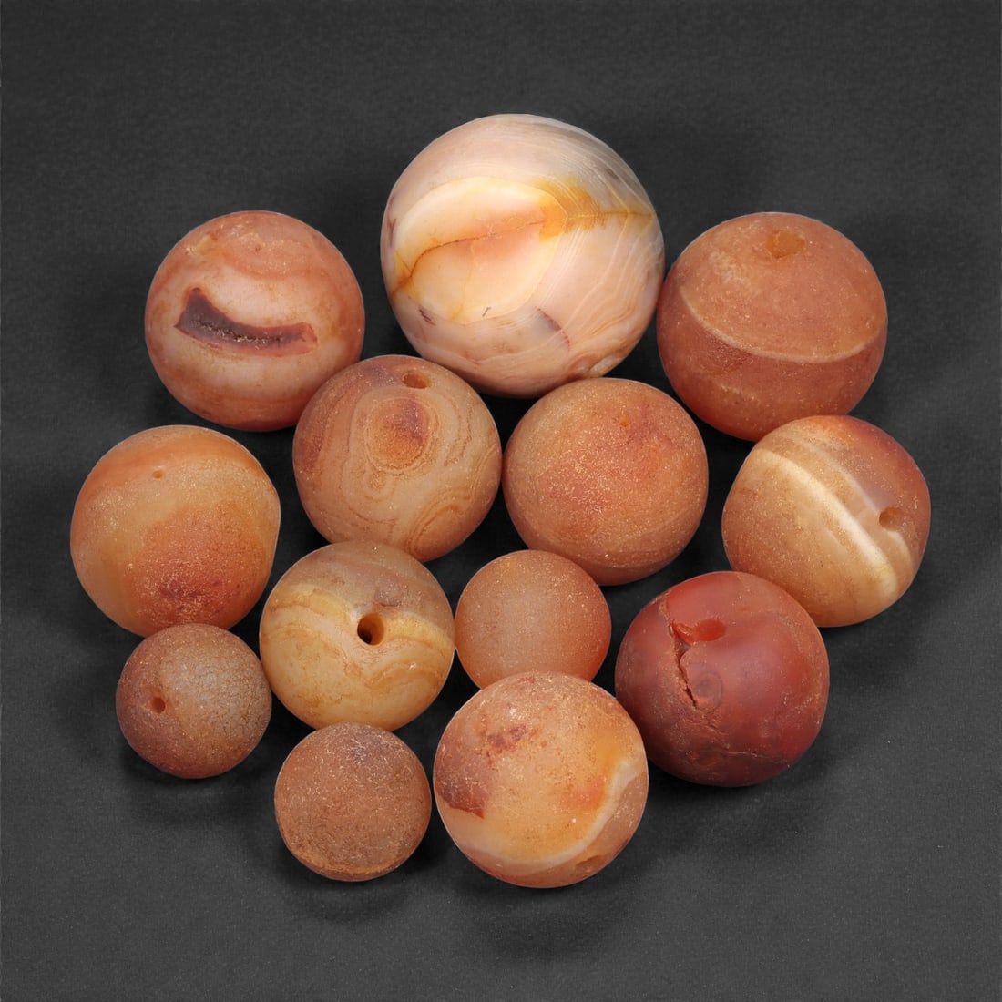 A Group of Tibetan Carnelian Beads, Early 20th Century, 1-2.2cm, 76LH: Comprising a group of spherical beads carved from orange to reddish-brown carnelian with natural banding and surface variation. Each is drilled longitudinally for stringing, consistent with traditiona