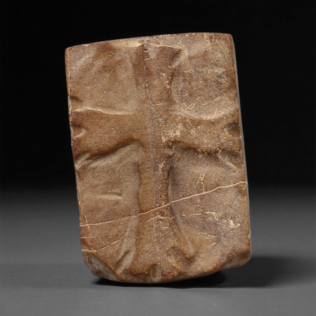A Late Antique Stone Relief with Cross Motif, 4th-6th Century CE, 20LA: Carved from a brown stone with natural surface veining, the relief is shaped as a rectangular plaque with a slightly rounded upper edge. The central field bears an incised cross with broad arms, defin