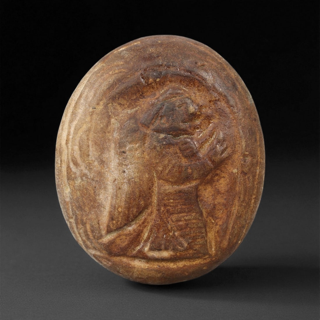A Byzantine Carved Stone Plaque, 6th-8th Century CE, 42LA: Carved from a brown stone with surface weathering and mineral accretions, the object is shaped as an oval plaque with a slightly convex face. The front bears an incised standing figure defined by line