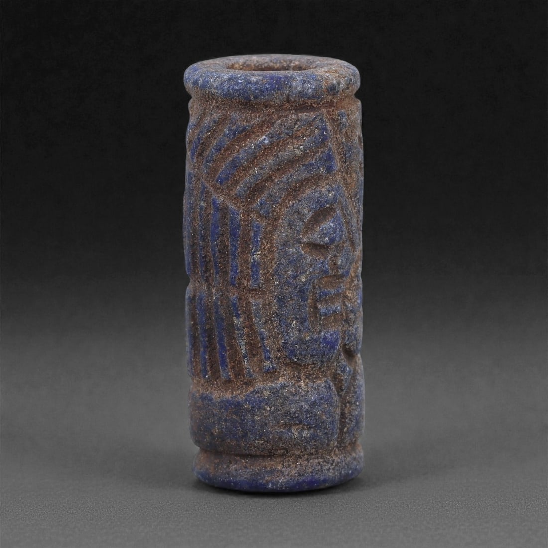 A Near Eastern Lapis Lazuli Cylindrical Relief Amulet, 3rd Millennium BCE, 3.1cm, 94L: Carved from lapis lazuli with mottled deep blue coloration interspersed with lighter calcite and brown mineral inclusions, the object is cylindrical in form with flat upper and lower faces. A longitud