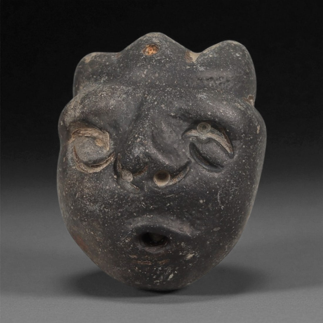 A Mediterranean Stone Maskette, 12th-17th Century, 89ZA: Carved from a dark grey stone with lighter surface abrasions, the maskette presents a rounded form tapering slightly toward the chin. The facial features are executed in shallow incised relief, includ