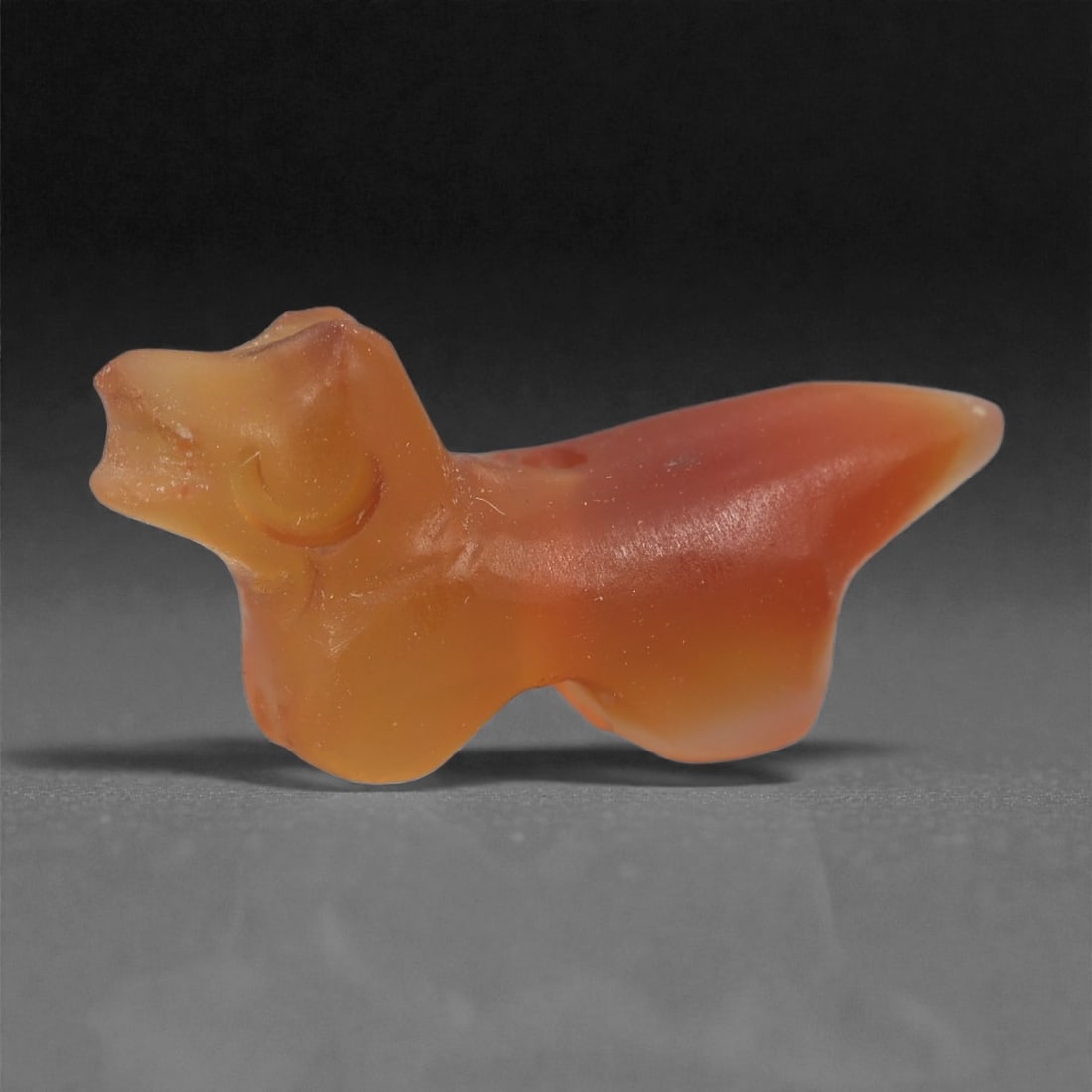 A Near Eastern Carnelian Zoomorphic Amulet, 1st Millennium BCE, 98LS: Carved from translucent orange carnelian, the amulet represents a small quadruped with an elongated body, short legs, and a raised head rendered with a simplified muzzle. The form is smoothly shaped w