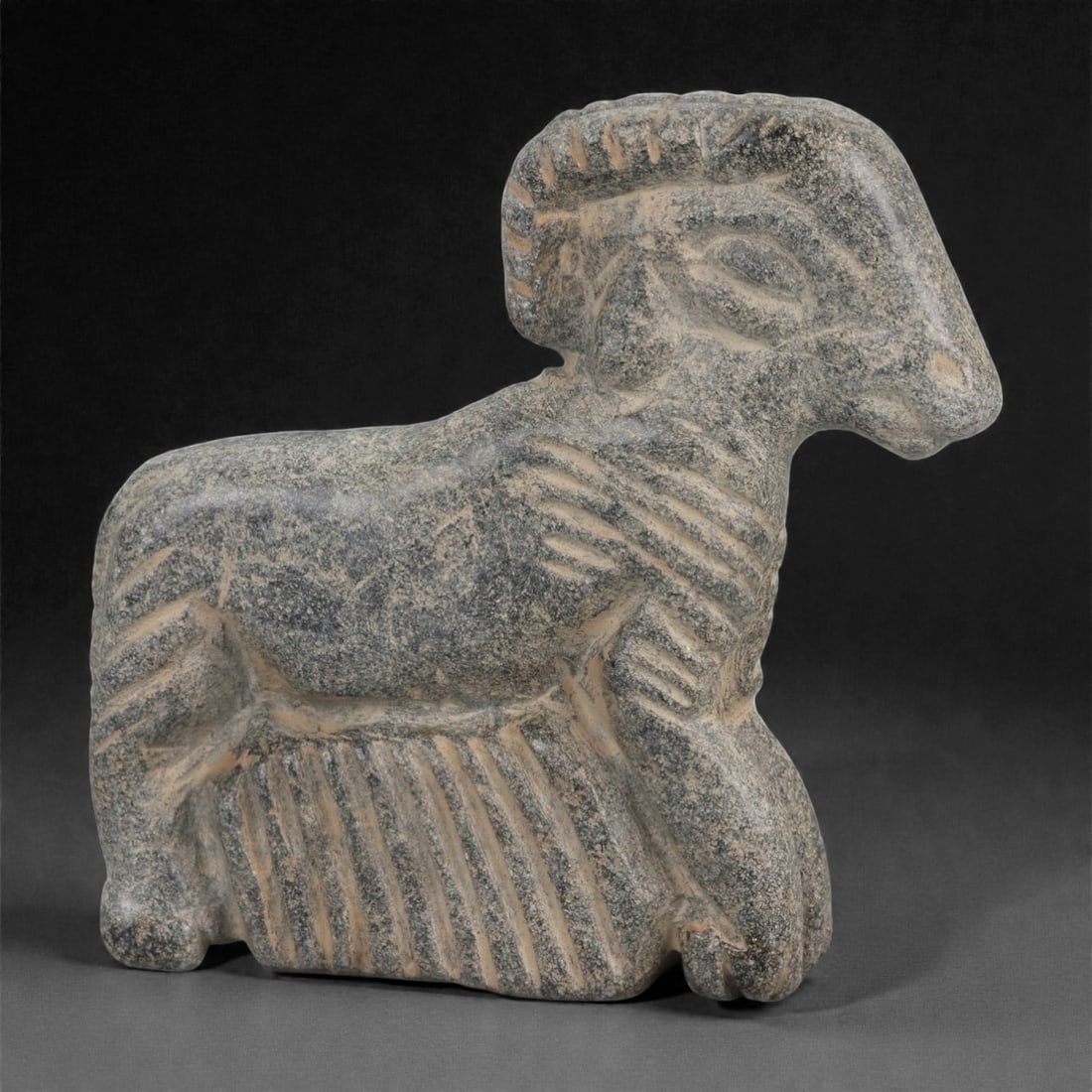 A Syro-Anatolian Schist Ram Effigy, Early to Mid-2nd Millennium BCE, 59LK: Carved from grey schist, the effigy depicts a ram in a seated posture with the forelegs drawn beneath the body and the hindquarters resting on a flat base. The head is elongated with a tapering muzzle