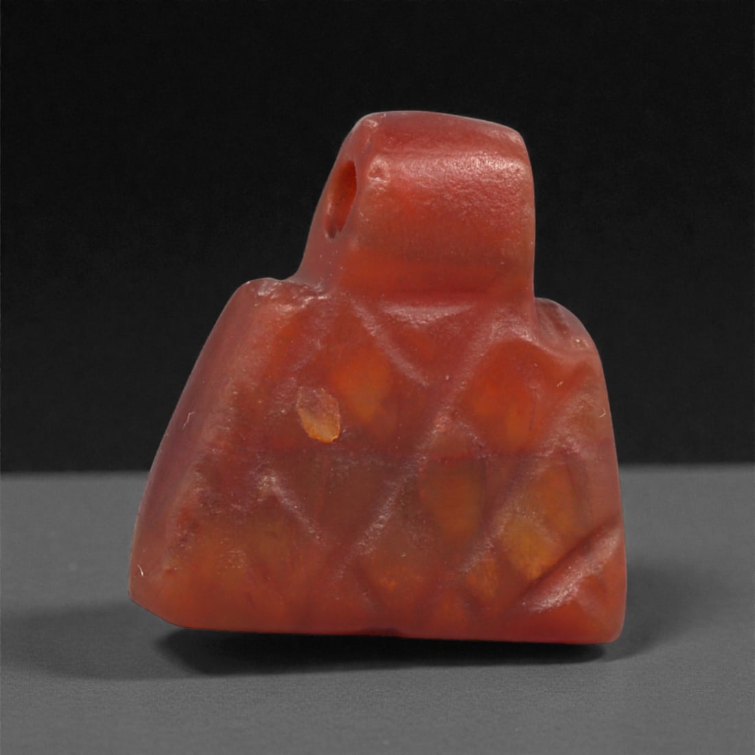 An Egyptian Carnelian Amulet, Coptic Period, 4th-7th Century CE, 1.4cm, 29LK: Worked from orange carnelian with natural internal variation, the amulet shows a compact form with smoothed contours and modest surface definition. Light wear is visible along the edges. Objects of th