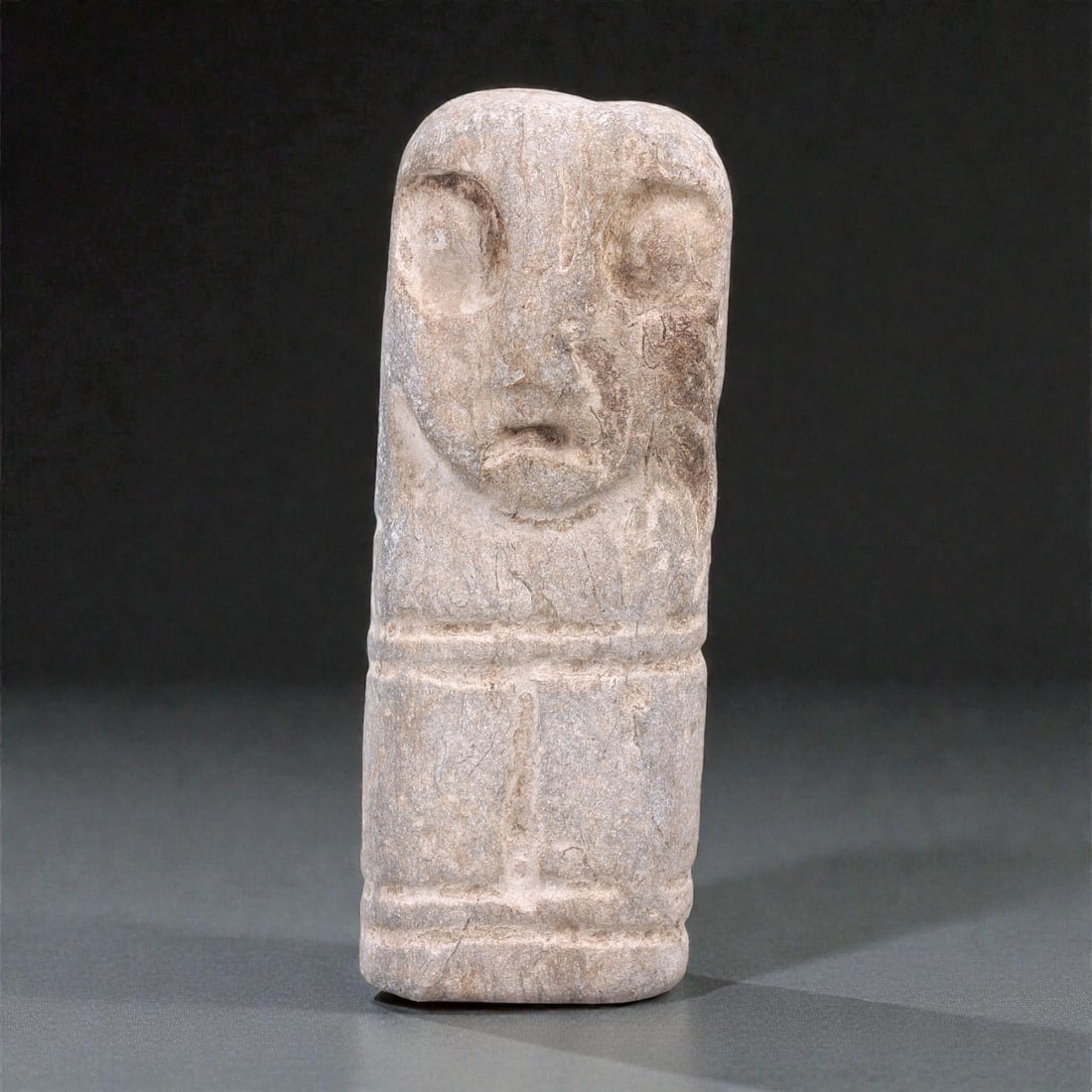 A Near Eastern Stone Idol, 4th-3rd Millennium BCE, 85R: Carved from light grey stone with natural inclusions and surface abrasion, the object takes the form of a vertically oriented cylindrical idol surmounted by a schematic human head. The facial features