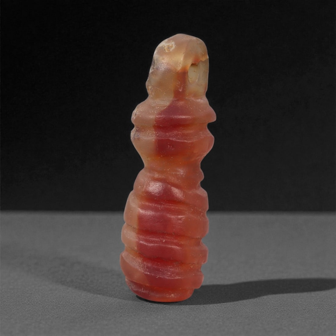 An Egyptian Carnelian Amulet, Coptic Period, 4th-7th Century CE, 2.6cm, 57LN: Worked from orange carnelian with natural internal variation, the amulet shows a compact form with smoothed contours and modest surface definition. Light wear is visible along the edges. Objects of th