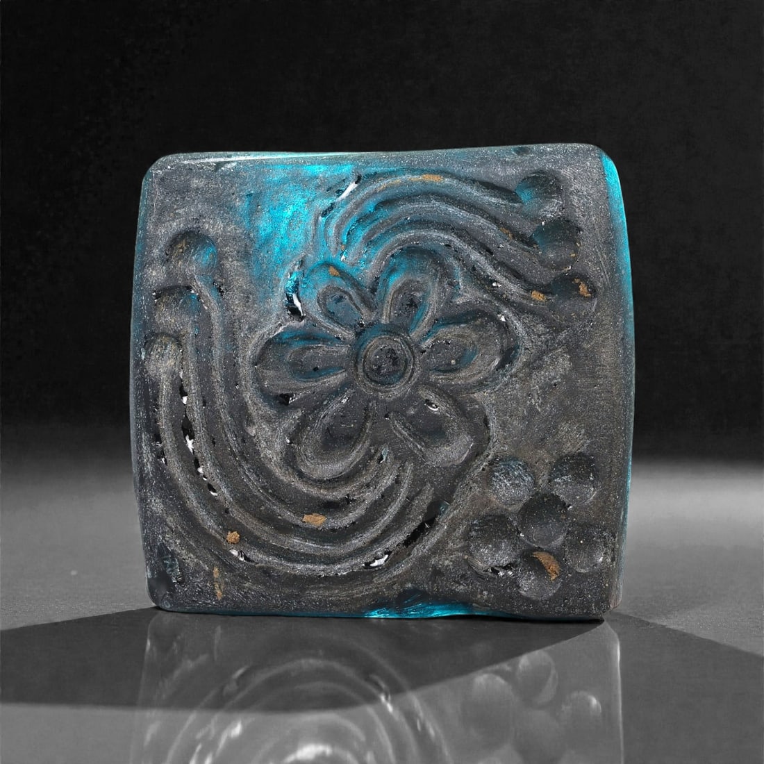 An Early Islamic Molded Blue Glass Plaque with Rosette, 8th-10th Century CE, 22LP: Cast in deep blue opaque glass with a matte, weathered surface, the plaque is rectangular with gently rounded corners. The obverse is molded in relief with a central six-petaled rosette framed by curv