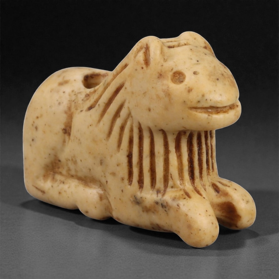 A Parthian Alabaster Feline Amulet, 1st Millennium CE, 13LA: Carved from a pale alabaster with scattered surface inclusions, the amulet depicts a recumbent feline with the forelegs extended and the hindquarters rounded. The head is modeled with a short muzzle, 