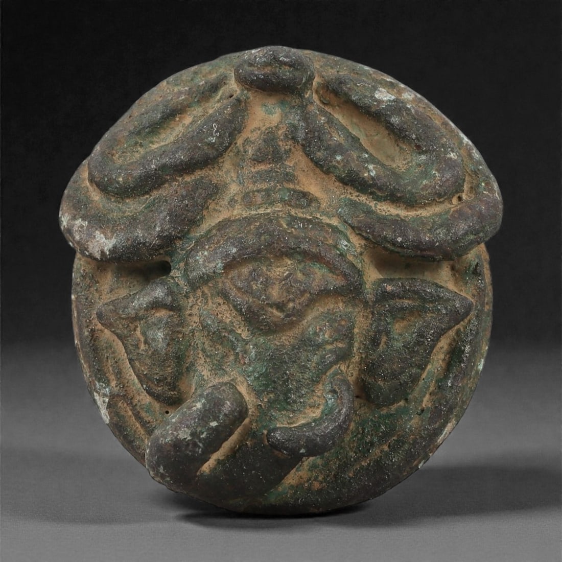 A Khmer Copper Alloy Elephant-Head Medallion, Angkor Period, 12th-13th Century, 6LS: Cast in copper alloy with a green patinated surface and areas of earthen accretion, the circular medallion bears a raised elephant head in frontal view, the broad ears flanking a central ridged forehe