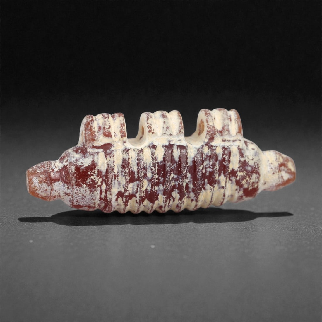 An Achaemenid Carnelian Amulet, 5th-4th Century BCE, 10A: Carved from deep red carnelian with areas of light surface accretion, the amulet is shaped as a cylindrical form with ribbed transverse grooves along the body. Each end tapers to a short conical termi