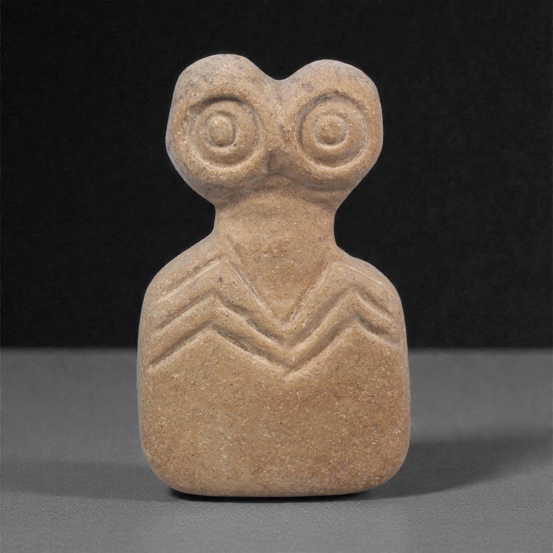 A Northern Mesopotamian Stone Eye Idol, 4AC: Carved from a fine-grained light brown stone, the figure presents a rectangular body surmounted by a bifurcated head with two recessed concentric circular eyes. The neck is subtly defined, and the upp